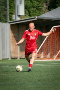 Soccer player in red uniform kicking ball on a green field during the day.